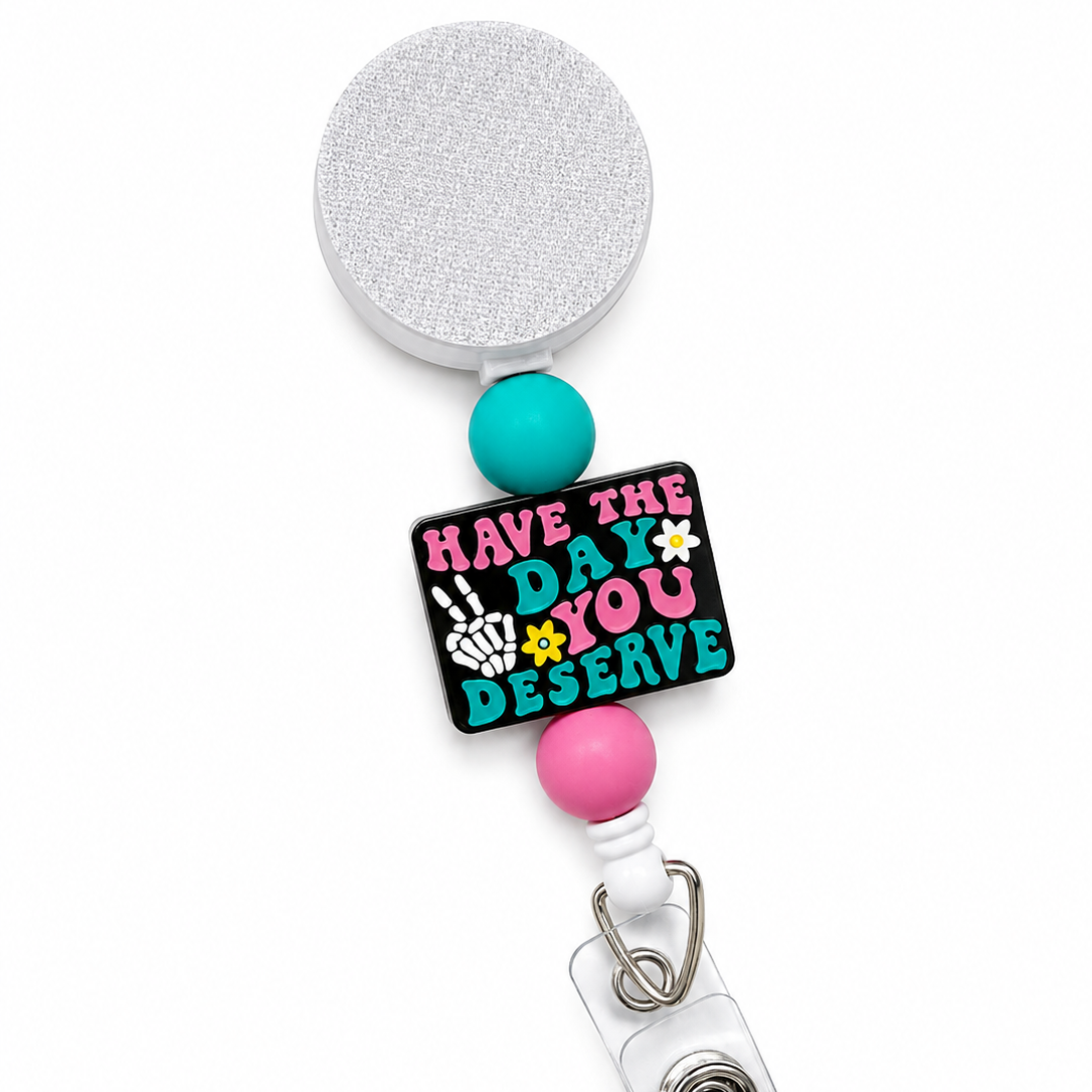 Have The Day You Deserve badge reel featuring a black statement topper with pink and teal lettering, skeleton hand peace sign, daisy accents, and teal and pink decorative beads on a white retractable badge reel.
