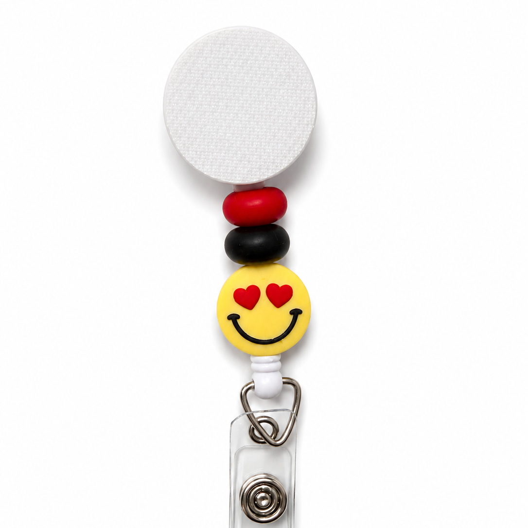 Heart eyes smiley face badge reel featuring a yellow emoji with red heart eyes, red and black decorative beads, and a white retractable badge reel. Cute work badge accessory.