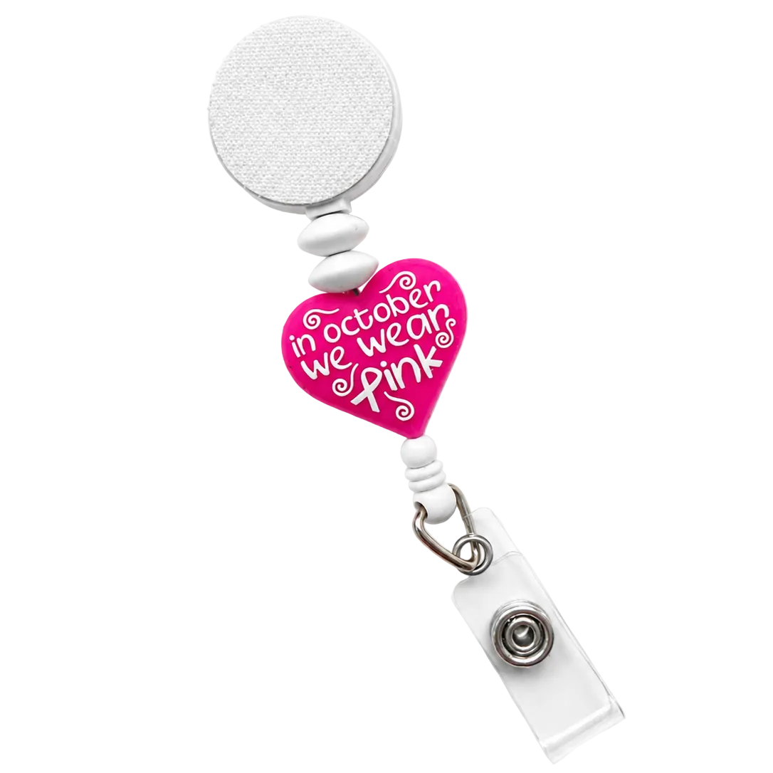 In October We Wear Pink badge reel featuring a bright pink heart with the words "In October We Wear Pink," white decorative beads, a retractable badge reel, and secure badge holder attachment.