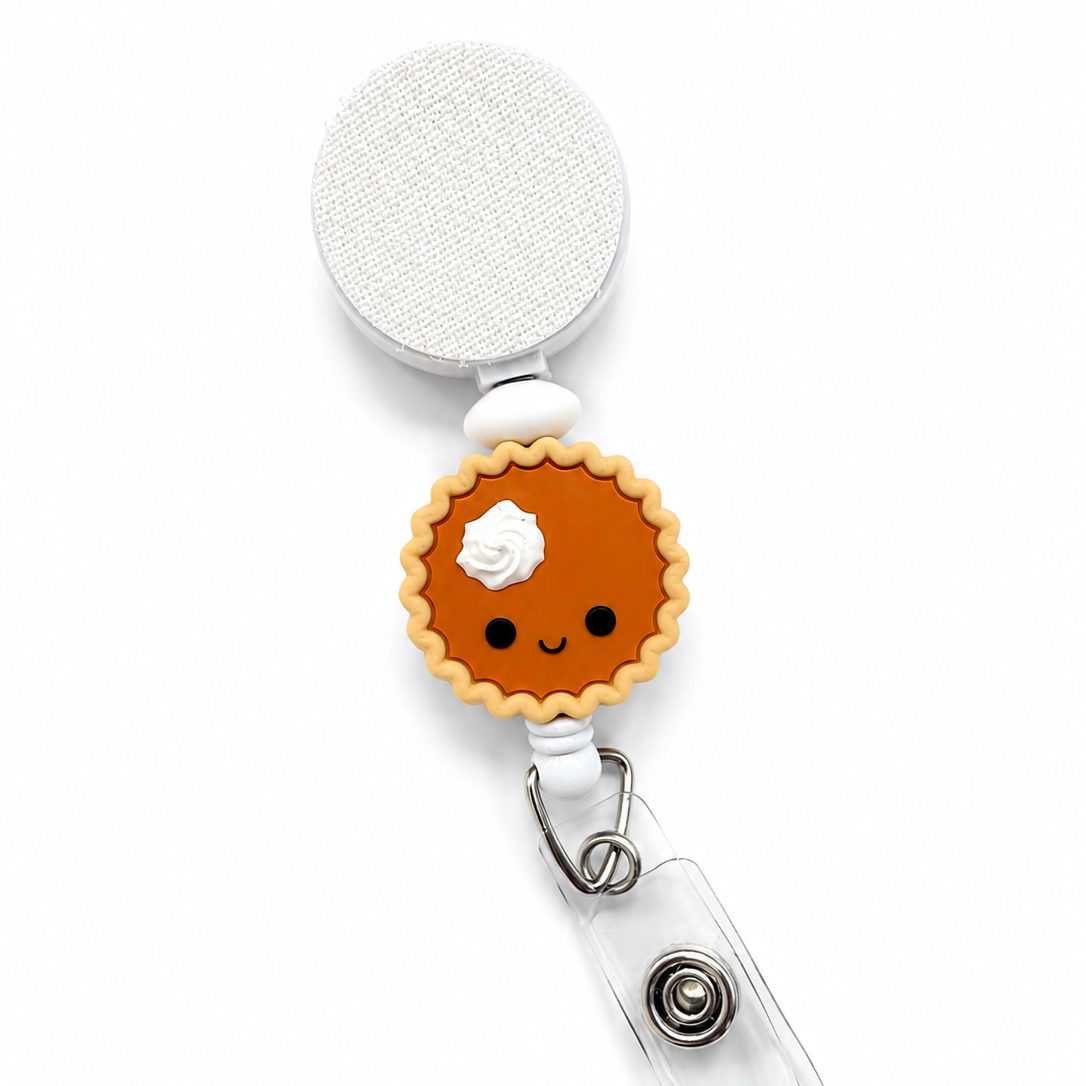Pumpkin pie badge reel featuring a smiling pumpkin pie with whipped cream on a white retractable badge reel. Cute Thanksgiving work badge accessory for fall.