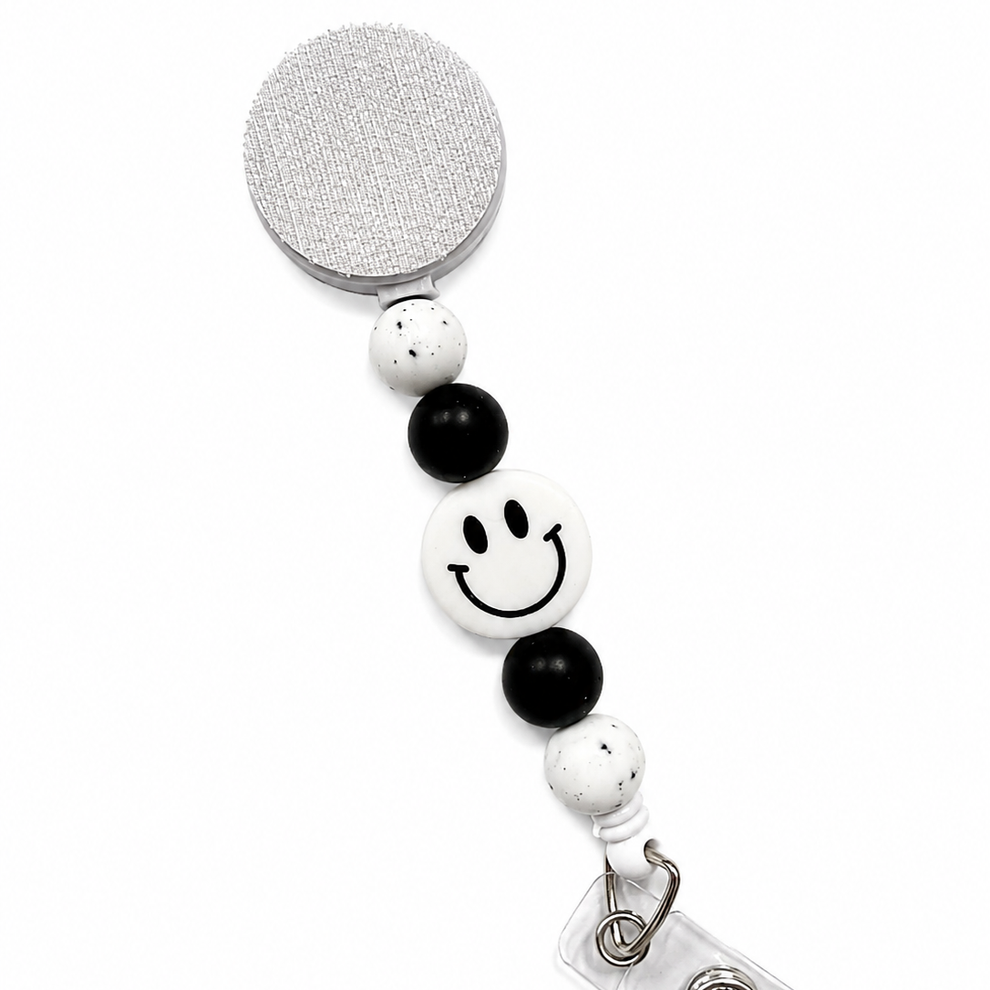 White Smiley Face badge reel featuring a white happy face design with black beads and white speckled decorative beads, retractable badge reel functionality, and secure badge holder attachment.